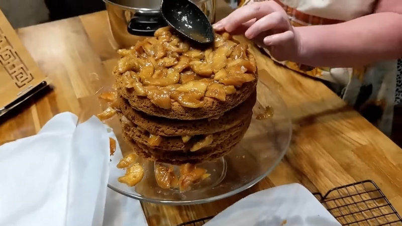A person spreads cooked apple filling on top of a layered apple stack cake using a spoon